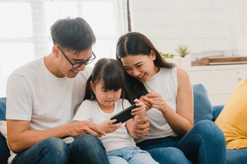 Happy Asian family dad, mom and daughter playing funny game online on smartphone sitting sofa in...