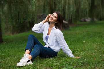 Fototapeta premium Attractive, happy and stylish brunette Caucasian girl posing while sitting on green grass in the park. Lifestyle and fashion concept.