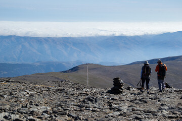 two hikers walking on slate ground descending from the top of Mulhacen, then a plain crossed by a path, then a mountain range and in the background a sea of low clouds