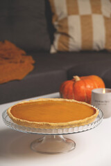 Pumpkin pie with Pumpkin and Concrete candlestick on table in living room next to sofa with pillows and blanket. Autumn comfort hyggelig at home. Background with copy space.