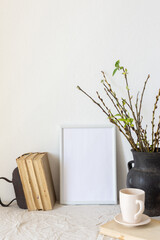 Home decor mock-up, blank picture frame near white painted concrete wall , black earthenware jug with pussy willow branches and blossoming green leaves, old books and a cup of coffee