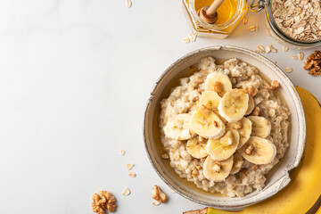 Oatmeal porridge in a bowl with banana, walnuts and honey on white background. Top view. Healthy...