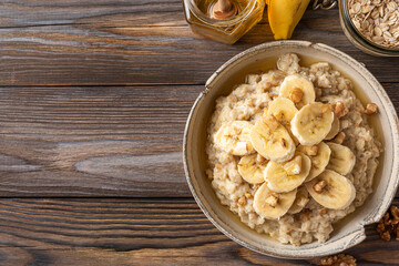 Banana, walnuts and honey oatmeal porridge in a bowl on rustic wooden table for healthy tasty breakfast. Top view
