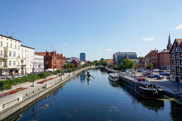 view of the historic waterfront warehouses and buildings on the Brda River in downtown Bygdoszcz with the Crossing the River sculpture in the foreground - obrazy, fototapety, plakaty