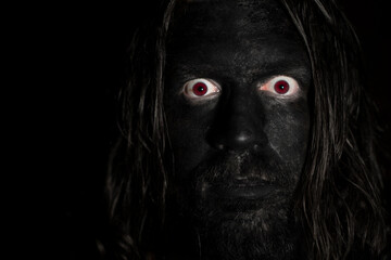 A scary face of a man in the dark. Long hair, black face and red eyes. Low light with dark background. Horror concept.