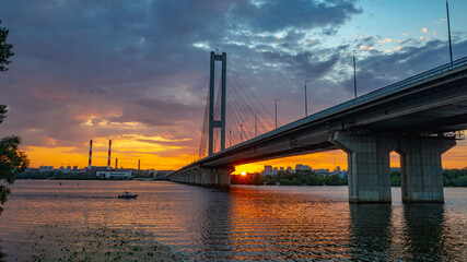 South bridge in Kiev. Sunset over the Dnieper. Thick clouds over the evening city. Evening shot of the bridge. Orange sun at sunset. The rays break through the clouds and are reflected in the river.