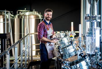 Young male brewer in leather apron supervising the process of beer fermentation at modern brewery...