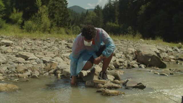 Professional black male scientist biologist in protective workwear analyzing biohazard danger and controlling environmental contamination, sampling polluted mountain river water into test tube.