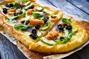 Pizza with smoked salmon, arugula, avocado, mozzarella and black olives on wooden background
