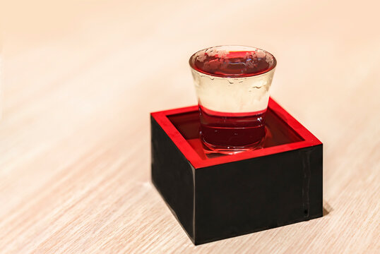 Chilled Japanese rice wine called nihonshu in a transparent overflowing sake-serving shot glass inside a traditional square lacquered wooden box called masu filled to the rim as a sign of prosperity.