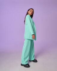 Elegant woman in mint costume blazer and pants and black top with stripes and boots standing and posing at purple background, Full length fashion portrait.