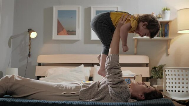 Mother and chils playing. Mom lies on bed and holds child in air, mother and baby playing and kiss, laugh. Slow motion