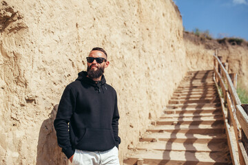 Young bearded hipster guy with beard in black blank hoodie or sweatshirt and sunglasses at the tropical beach. Mock-up for print.