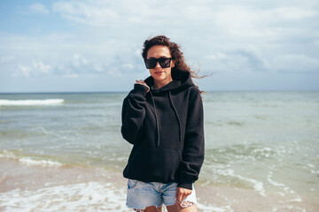 Beautiful young adult woman in black hoodie or sweatshirt and sunglasses at the tropical beach. Mock-up for print. Space for your logo or design.