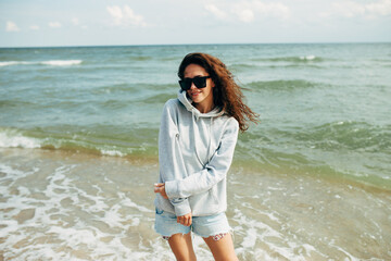 Beautiful young adult woman in gray blank hoodie or sweatshirt and sunglasses at the tropical beach. Mock-up for print. Space for your logo or design.