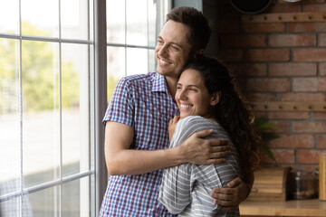 Dreamy loving young husband cuddling happy beautiful hispanic wife, visualizing future looking in distance out of window, enjoying tender sweet family moment or celebrating moving into own house.