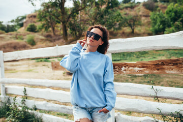 Beautiful young adult woman in blue blank sweatshirt and sunglasses on the background of a wooden fence in nature. Mock-up for print. Space for your logo or design.