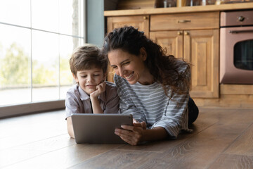 Happy young hispanic mother using digital tablet with laughing little preschool kid son, lying together on warm wooden floor in kitchen, watching funny cartoons or video content in social network.