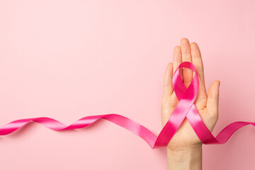 First person top view photo of female hand holding pink ribbon in palm symbol of breast cancer awareness on isolated pastel pink background with copyspace