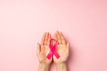 First person top view photo of girl's hands holding pink ribbon in palms symbol of breast cancer awareness on isolated pastel pink background with blank space