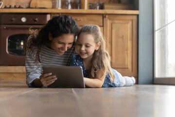 Happy young mother watching cartoons or funny photo video content in social networks on tablet with cute smiling little kid daughter, lying together on heated warm floor, tech addiction concept.