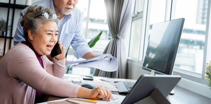 Elderly senior asian couple male female freelancer in casual clothes typing on laptop keyboard while talking on smartphone sitting at desk and busy working in home office - Powered by Adobe
