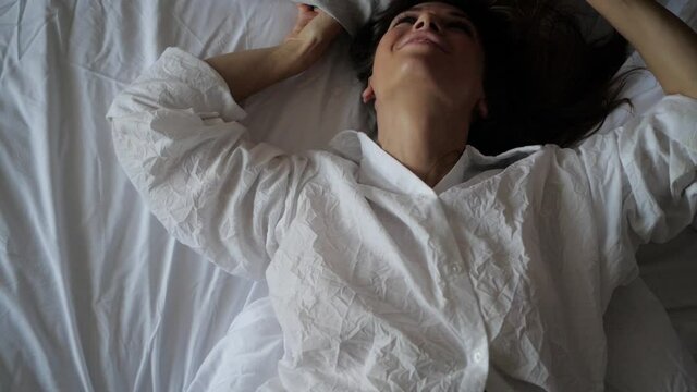 a brunette woman in a white shirt lies on a white sheet and throws up a pillow, smiles at the camera. High quality FullHD footage