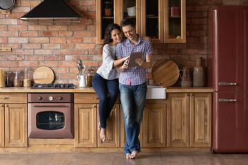 Full length relaxed smiling young family couple spending time in stylish wooden kitchen, using digital computer tablet, choosing goods shopping in internet store, web surfing or playing online.
