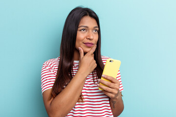 Young Venezuelan woman holding mobile phone isolated on blue background looking sideways with...