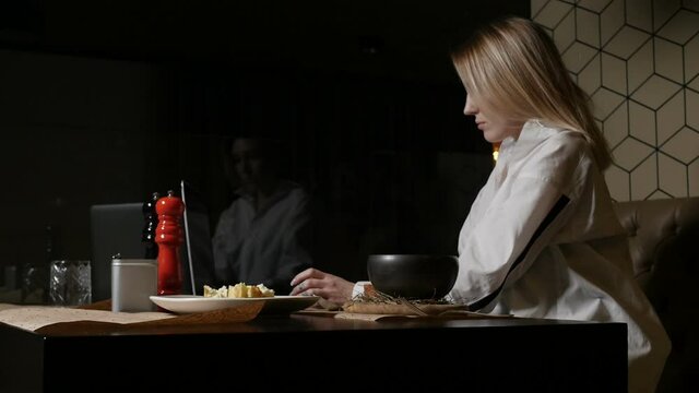 blonde girl in a white shirt eats a dish sitting on a brown sofa in a restaurant. High quality FullHD footage