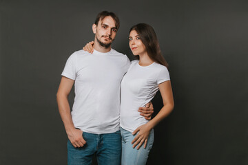 Fototapeta premium A young couple, a girl and a man in white blank t-shirts on a dark gray background. Mock-up. love story