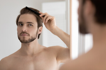 Handsome shirtless bearded 30s guy standing in bathroom after shower looks in mirror holding wooden...