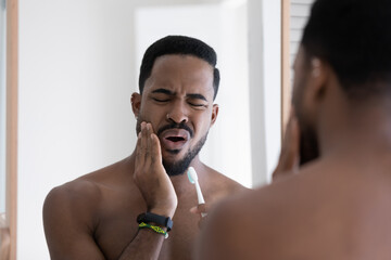 Handsome 30s African guy with frown face expression reflected in mirror touch cheek while brush...