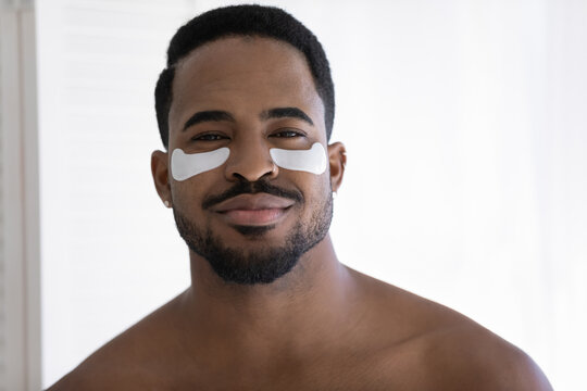 Head shot portrait of handsome African shirtless man with applied moisturizing under-eye anti-wrinkle patches pose look at camera. Facial beauty skincare, removing bags and circles treatment concept