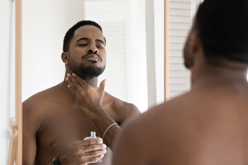 Handsome young African shirtless man reflected in mirror applying to neck perfume or to skin...