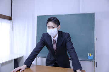 黒板の前でマスクをして立つスーツ姿の教師