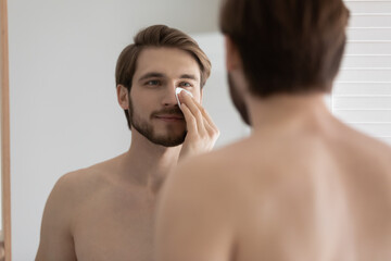 Young man look in mirror holds cotton pad cleaning face use cleansing tonic, apply aftershave lotion, removing facial dirt, daily morning routine, personal skincare, beauty treatment cosmetic concept