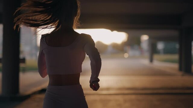 A young woman runner is training in the summer within a city. Close up of silhouette fitness girl is jogging outdoor. Concept of workouts running and healthy lifestyle. Back view. Slow motion.