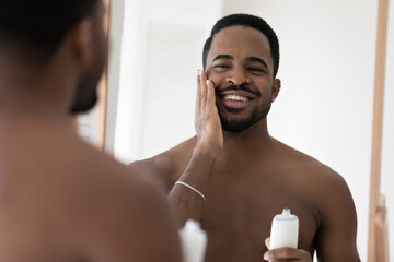 African shirtless man do morning self-hygiene routine holds bottle apply skincare cosmetics...