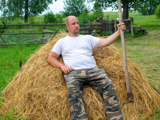 A middle-aged man dressed in trousers and a T-shirt is sitting on a stack of yellow hay and holding...