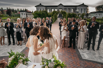 Portrait of two LGBT females lesbians brides kissing during wedding ceremony, guests clapping and...