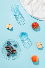 Creative composition made of plate with fruits, glasses, apples on pastel blue sunlit background with white silk cloth and palm tree leaves shadows. Summer and refreshment concept. Harvest theme