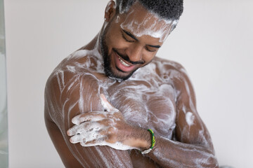Close up cropped view young African man lather his muscular build body with hydrating shower gel,...