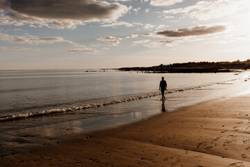 Naklejka premium Sunset on a beach in Ireland. A person is walking on the beach of Silverstrand near Galway. Golden hour beach image with clouds and a silhouette of a man. 