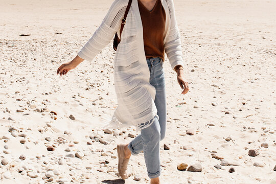 A woman is walking on a sandy beach. A woman in blue jeans and brown top with white cardigan is walking on a sandy beach in Ireland. 