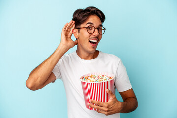 Young mixed race man eating popcorns isolated on blue background trying to listening a gossip.