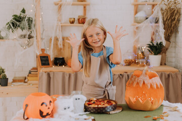a blonde baby girl with long hair in an apron in the kitchen, decorated with pumpkins and garlands for Halloween, prepares a focaccia pie. space for text. High quality photo
