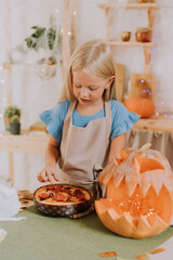 a blonde baby girl with long hair in an apron in the kitchen, decorated with pumpkins and garlands for Halloween, prepares a focaccia pie. space for text. High quality photo