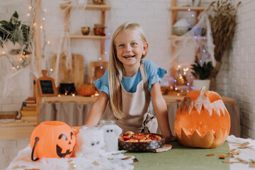 a blonde baby girl with long hair in an apron in the kitchen, decorated with pumpkins and garlands for Halloween, prepares a focaccia pie. space for text. High quality photo