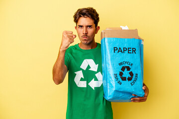 Young mixed race man recycling paper isolated on yellow background showing fist to camera, aggressive facial expression.
